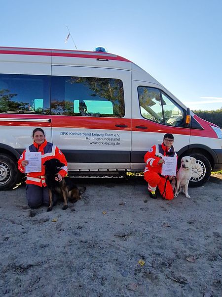 zwei Personen mit Rettungshunden posieren vor Fahrzeug nach bestandener Prüfung